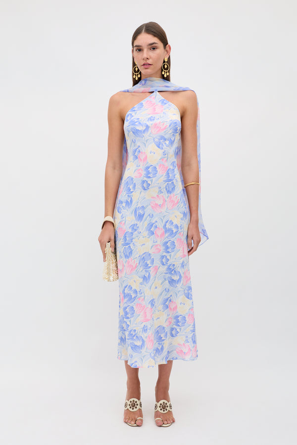 A woman stands against a plain background wearing the Hayley - Tulip Fields Blue dress, a pastel floral halter-neck style in silk crepe de chine with a detachable georgette scarf, styled with gold statement earrings, bracelet, heeled sandals, and a small beaded bag.
