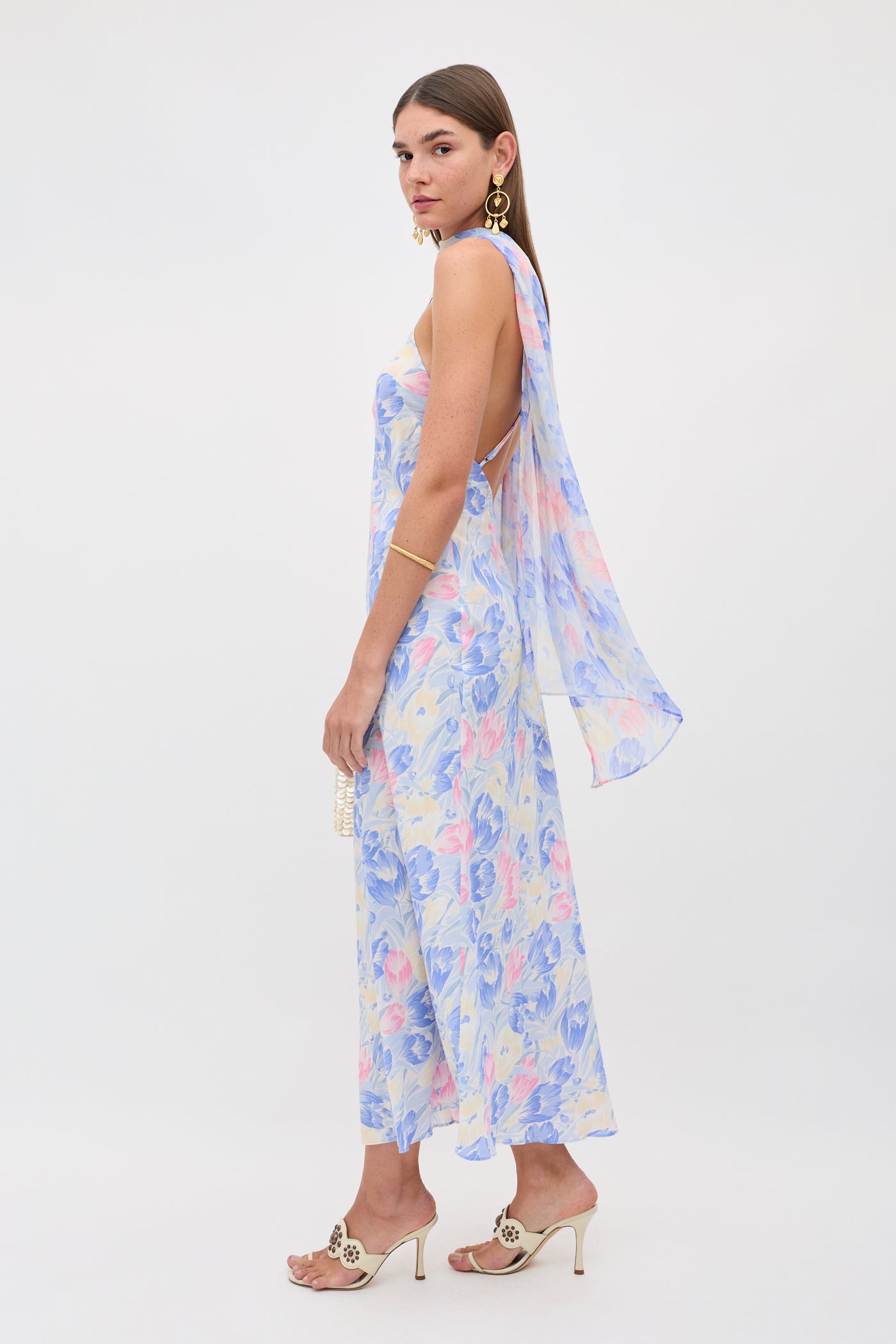 A woman stands sideways wearing the Hayley - Tulip Fields Blue—a flowy, sleeveless silk crepe de chine dress with a detachable georgette scarf—accessorized with gold jewelry, heeled sandals, and a small clutch against a plain white background.