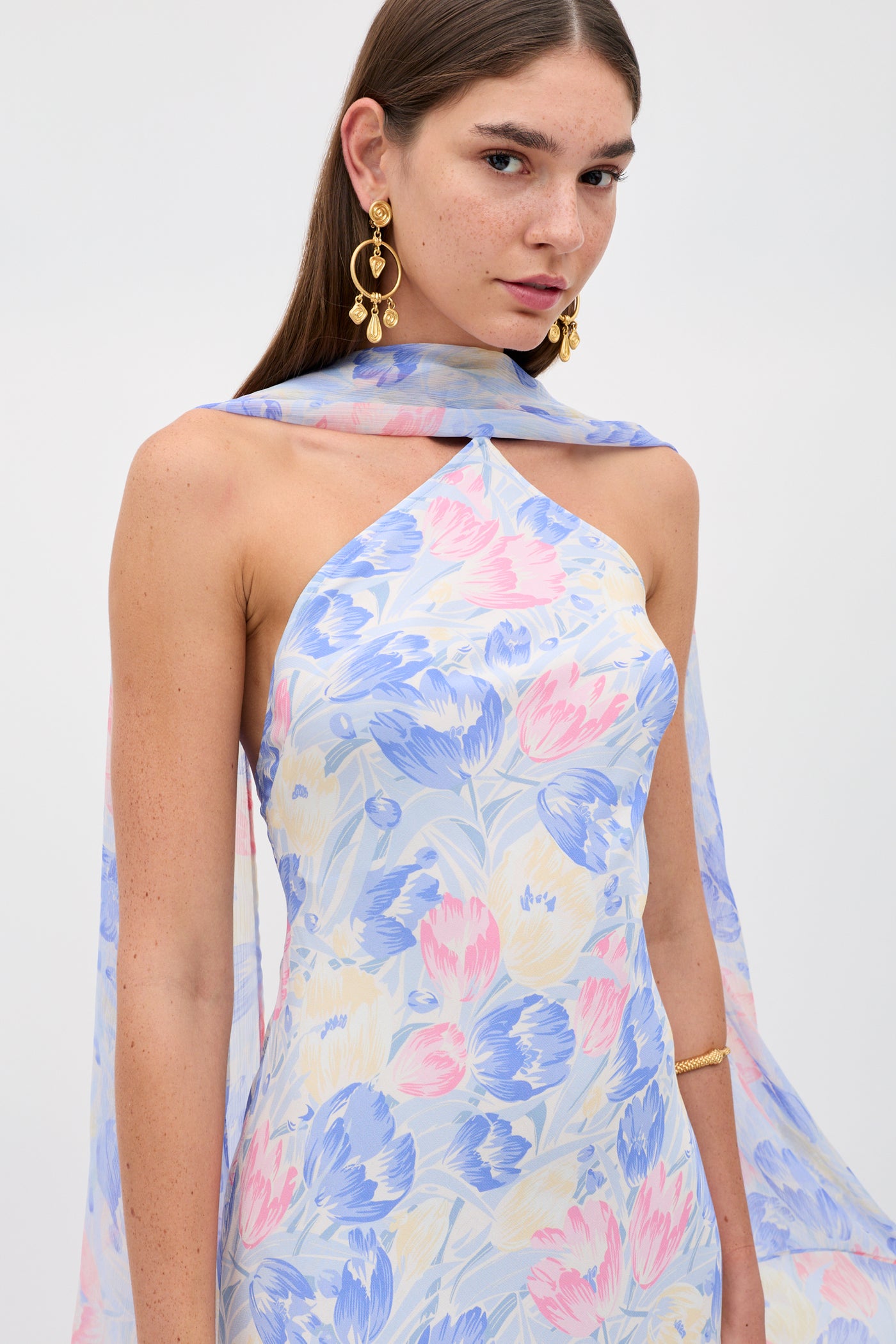 A woman wearing the Hayley - Tulip Fields Blue dress in silk crepe de chine with a detachable georgette scarf, paired with gold dangling earrings and a gold bracelet, stands against a plain white background.