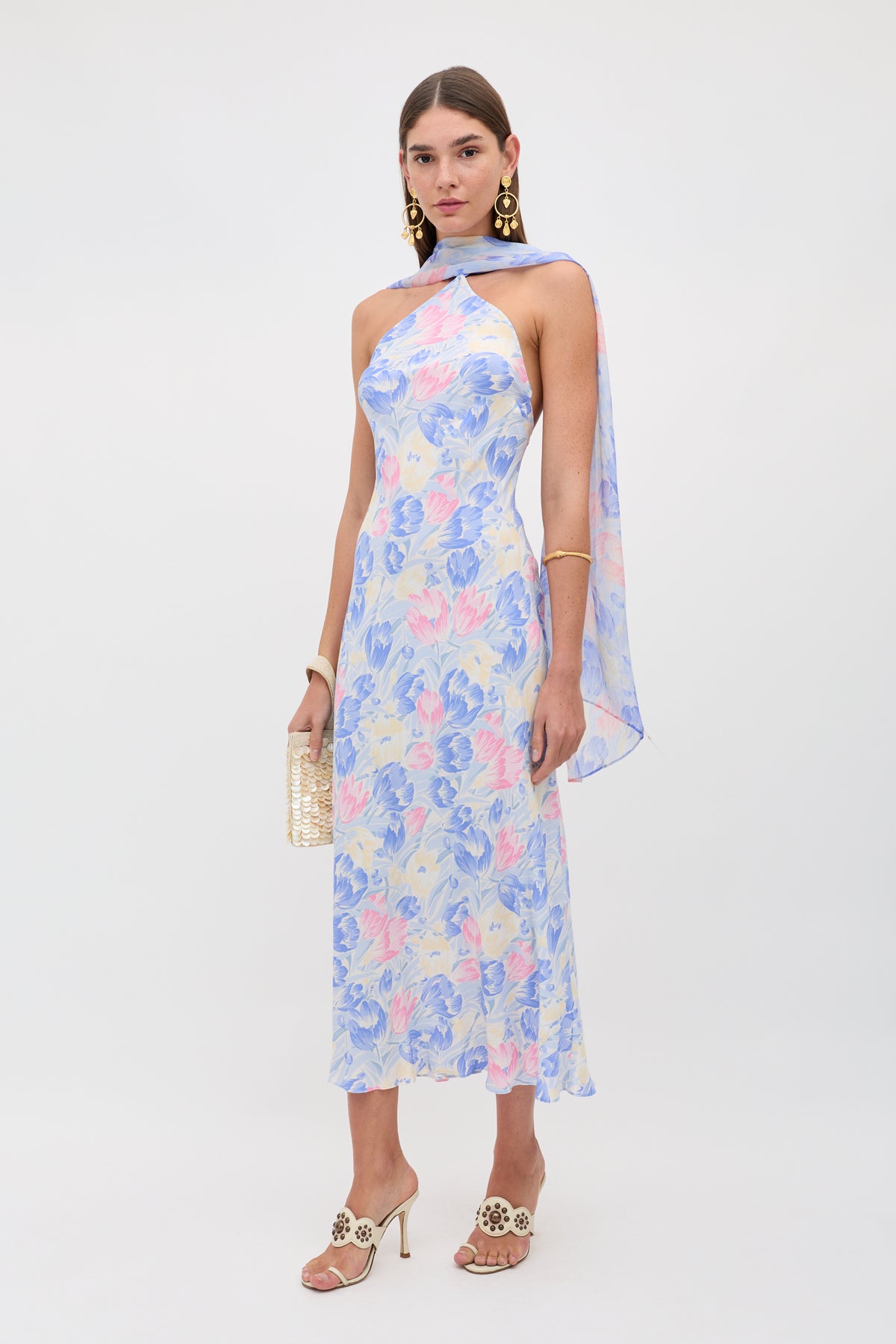 A woman wearing the Hayley - Tulip Fields Blue dress—a sleeveless, pastel floral silk crepe de chine midi with a detachable georgette scarf—plus gold earrings, beige heeled sandals, and a woven clutch stands against a plain white background.