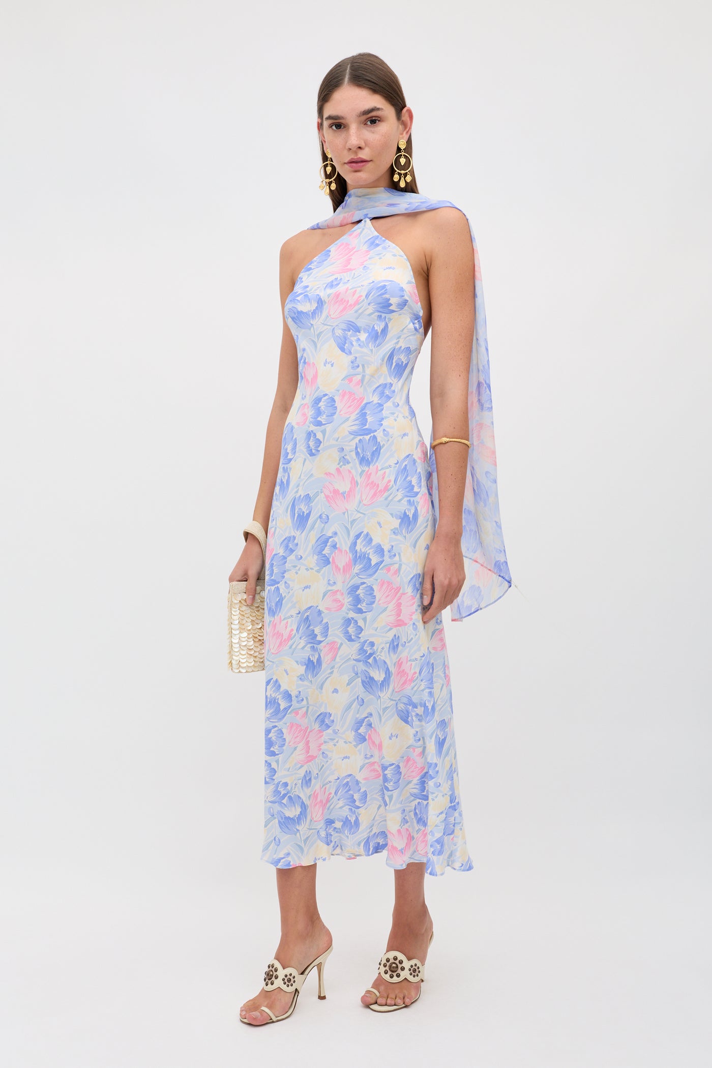 A woman wearing the Hayley - Tulip Fields Blue dress—a sleeveless, pastel floral silk crepe de chine midi with a detachable georgette scarf—plus gold earrings, beige heeled sandals, and a woven clutch stands against a plain white background.