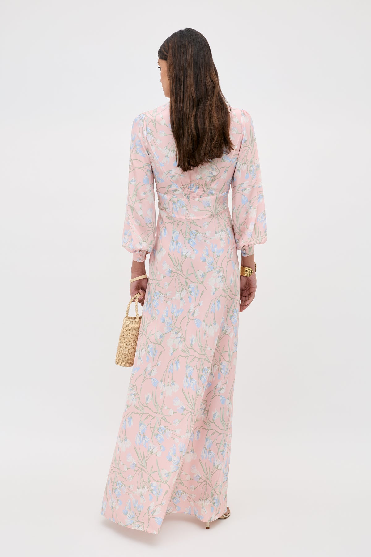 A woman with long brown hair faces away, wearing the Emory - Watercolour Floral Pink maxi dress with long sleeves. She carries a small woven handbag and sports a gold watch on her left wrist, set against a plain white background.