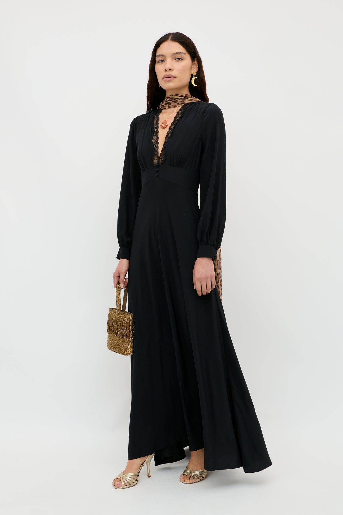 Wearing “Emory - Black,” a long, elegant dress crafted from silk crepe de chine with sheer lace details, the woman holds a woven handbag. She completes her sophisticated look with gold heels, a statement earring, and straight brown hair as she stands against a plain white background—perfectly styled for any special occasion.