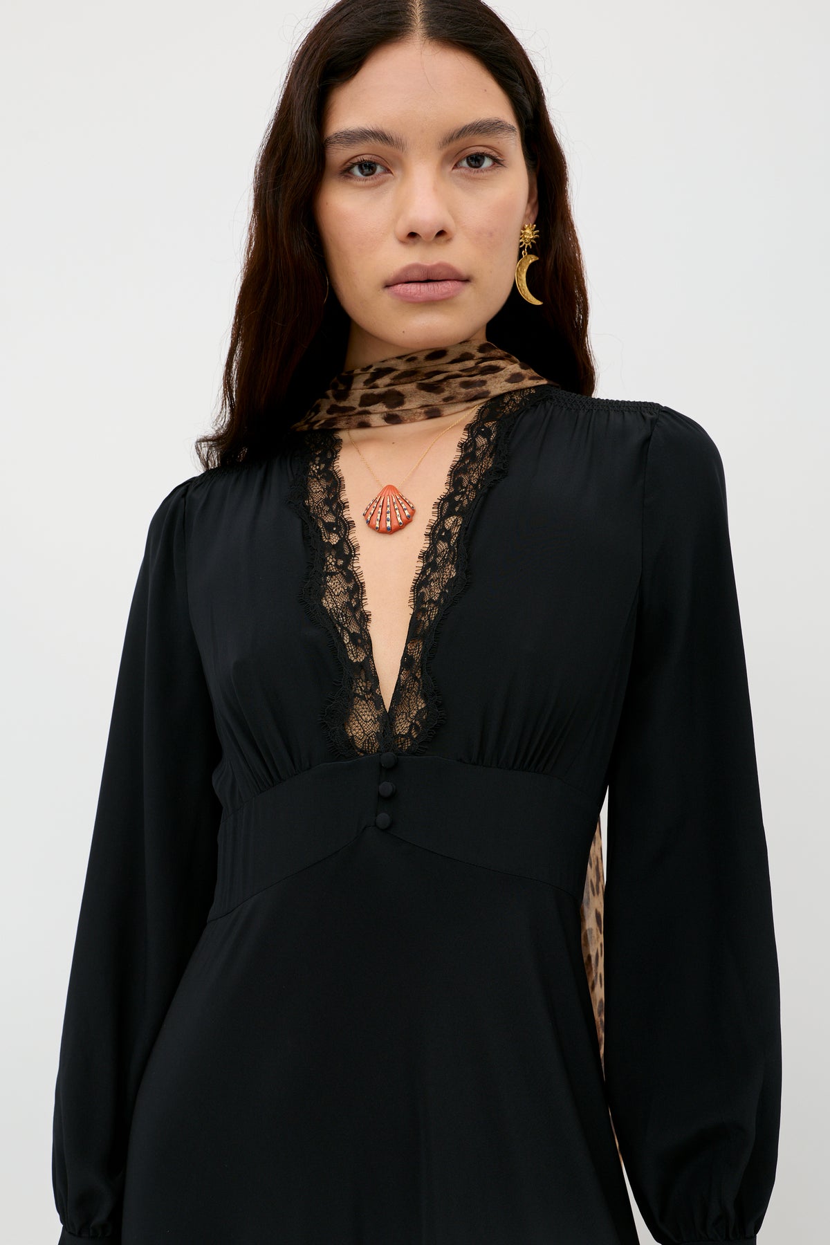 Wearing the Emory – Black, a silk crepe de chine dress with lace trim, the woman with long dark hair stands facing forward against a plain light background. She completes her elegant look with a leopard-print scarf, gold earring, and a red shell-shaped pendant—an ensemble perfect for special occasions.