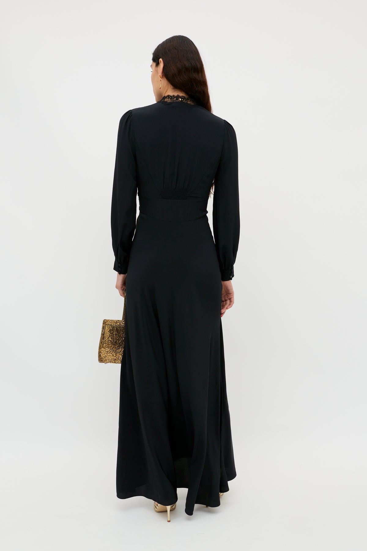 A woman with long dark hair, dressed in the elegant Emory – Black silk crepe de chine dress and holding a small textured gold handbag, stands facing away from the camera against a plain white background—perfect for a special occasion.