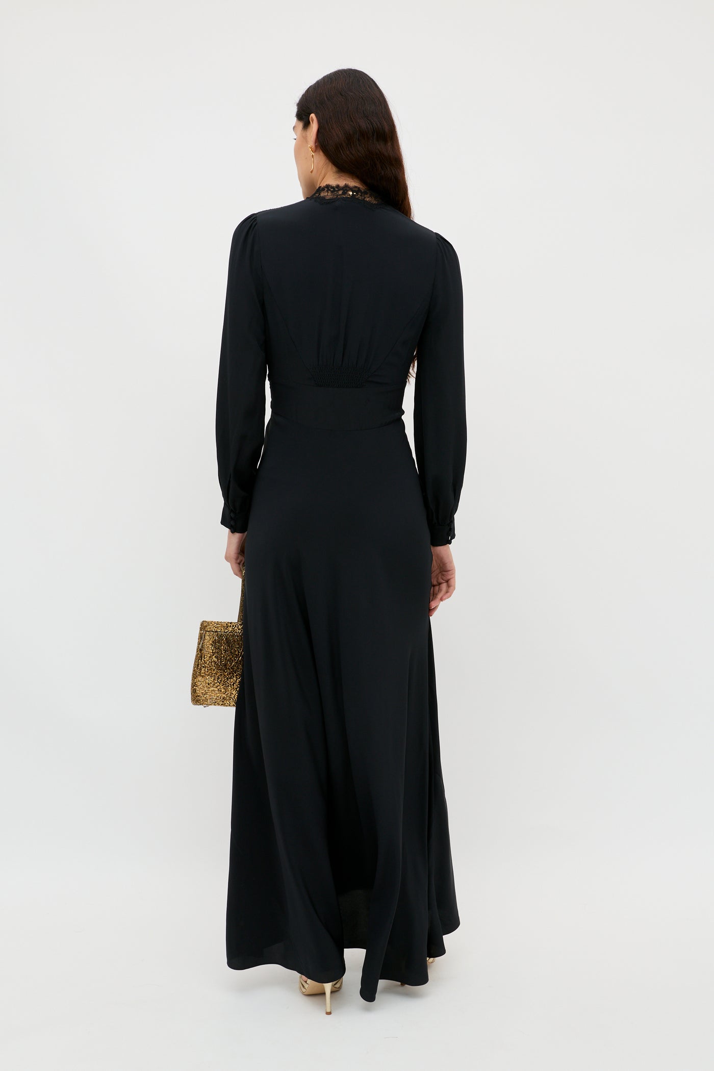 A woman with long dark hair, dressed in the elegant Emory – Black silk crepe de chine dress and holding a small textured gold handbag, stands facing away from the camera against a plain white background—perfect for a special occasion.