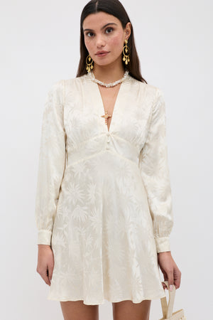 A woman with straight brown hair wears the Hebe - Daisy Jacquard Champagne mini dress, paired with gold earrings, a necklace, and holding a small handbag against a plain background.