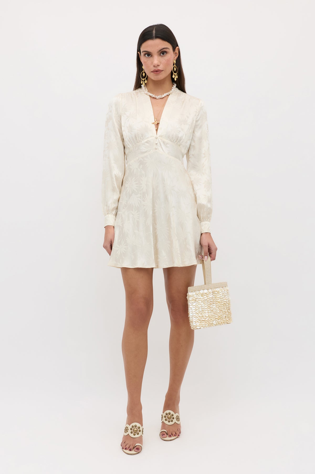 A woman stands against a white background wearing the Hebe - Daisy Jacquard Champagne mini dress with a v-neckline, white floral slide sandals, large gold earrings, and holding a small woven handbag.