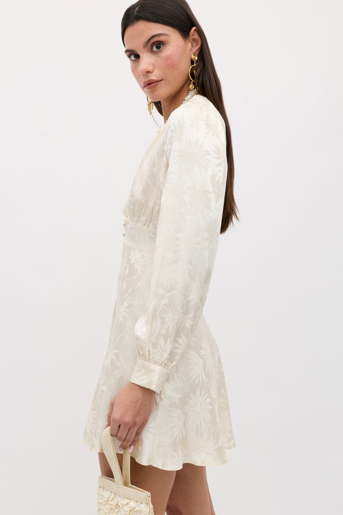 A woman with long dark hair and gold earrings wears the Hebe - Daisy Jacquard Champagne dress, featuring cream floral jacquard, as she holds a small textured bag and looks over her shoulder against a plain light background.