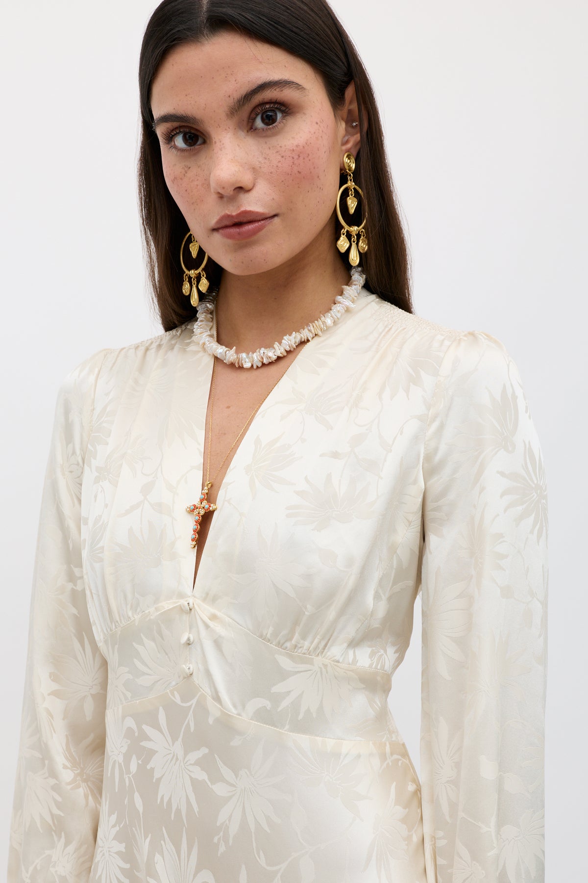 A woman with long brown hair wears the Hebe - Daisy Jacquard Champagne dress with long sleeves, styled with gold earrings and a white beaded necklace. She has subtle makeup and is set against a plain white background.