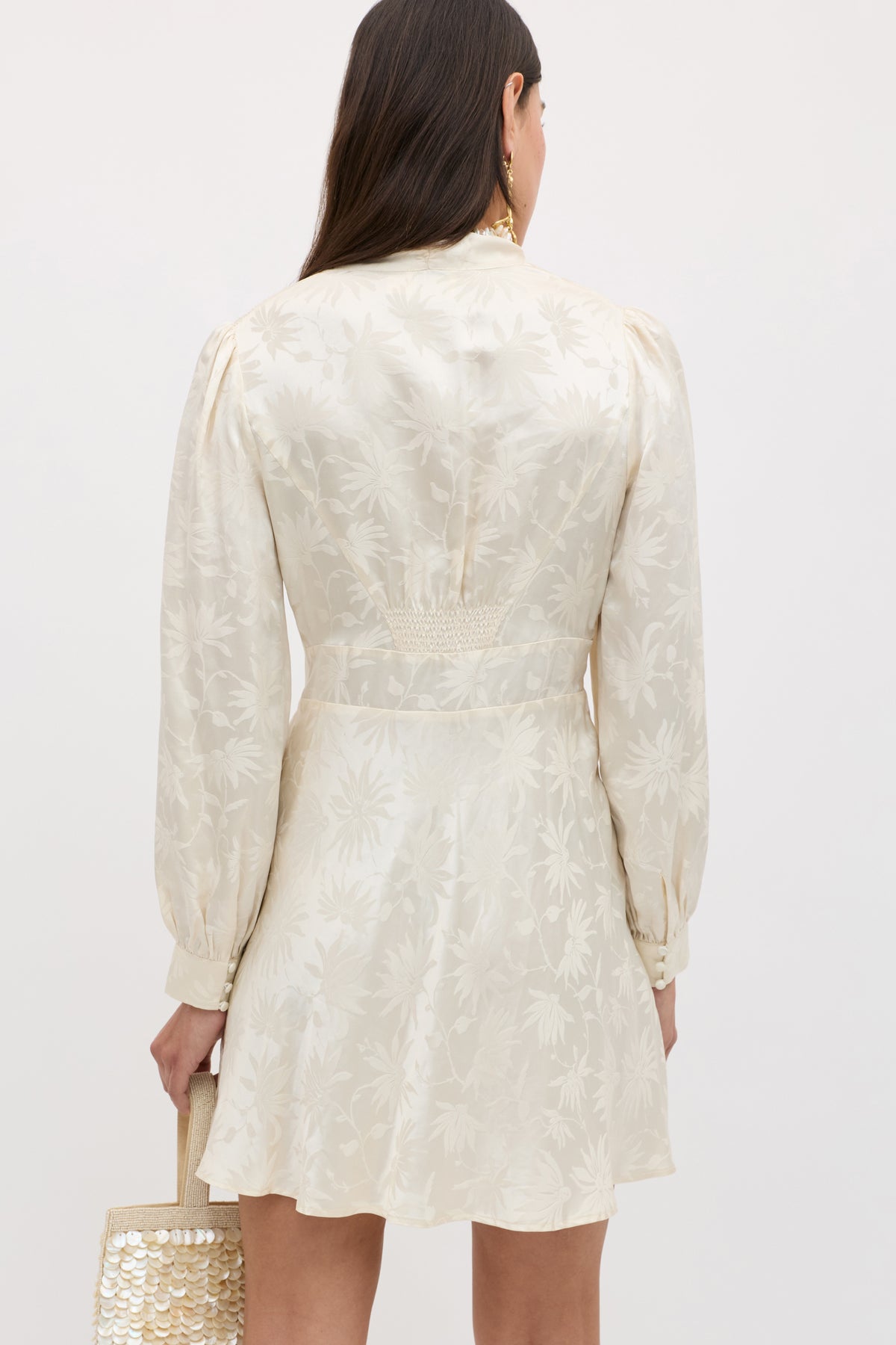 Seen from the back, a woman with long brown hair wears the Hebe - Daisy Jacquard Champagne, a cream-colored silk mini dress with jacquard daisies, and carries a small light beige sequined handbag.