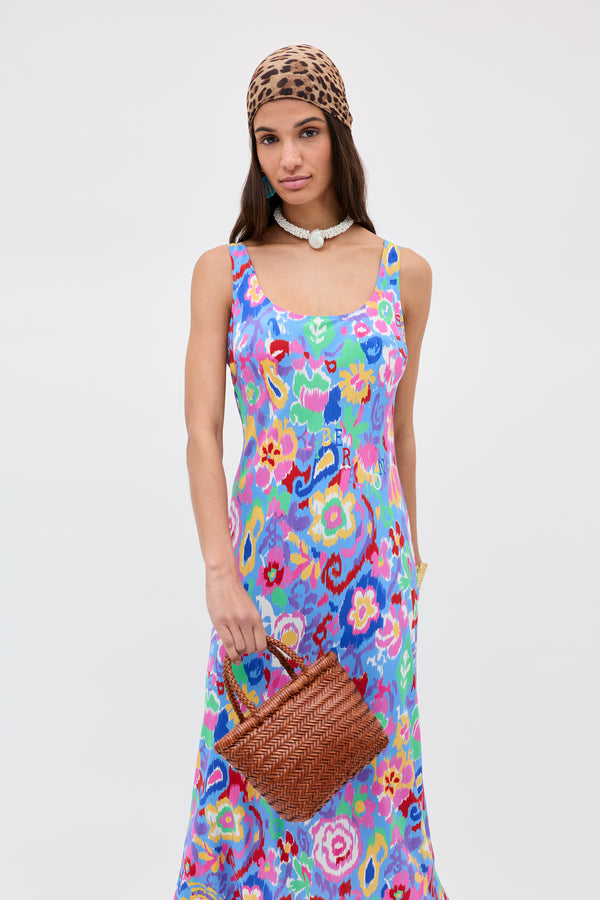 A woman in a colorful floral dress, leopard print headscarf, and white beaded necklace holds the Lysia - Tan woven bag—showcasing craftsmanship reminiscent of Dragon Diffusion—against a plain light background.