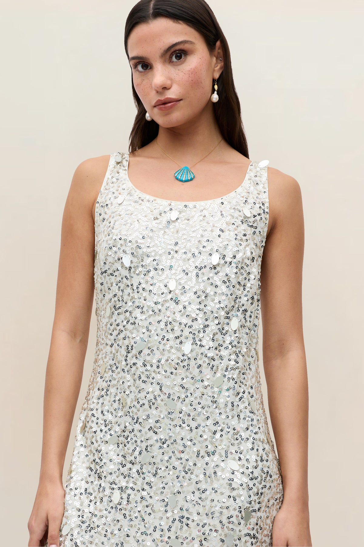A woman wearing the Bondi Mia Squiggle Embellishment Ivory dress stands against a plain background. She has long brown hair and is accessorized with pearl earrings and a blue shell-shaped necklace.