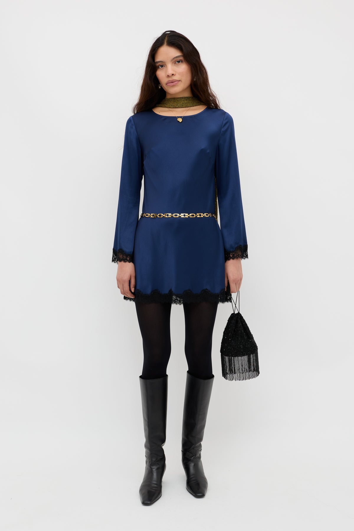 A woman stands against a plain white background, wearing the Ridley - Midnight Navy long-sleeved dress with black lace trim, paired with black tights, knee-high boots, and holding a small fringed black bag.