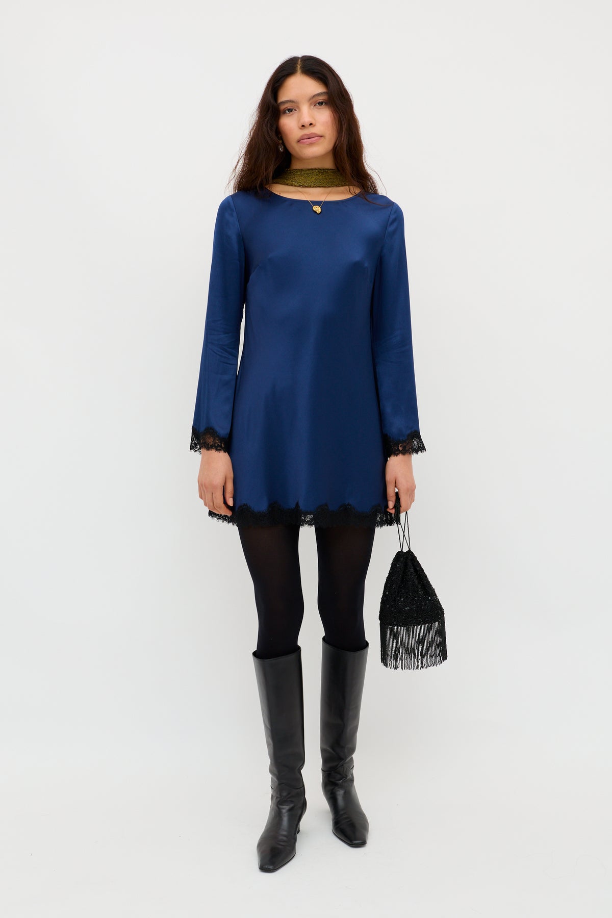 A woman stands against a white background wearing the Ridley - Midnight Navy long-sleeve mini dress with black lace trim, paired with black tights, knee-high black boots, a small fringed bag, and loose hair.