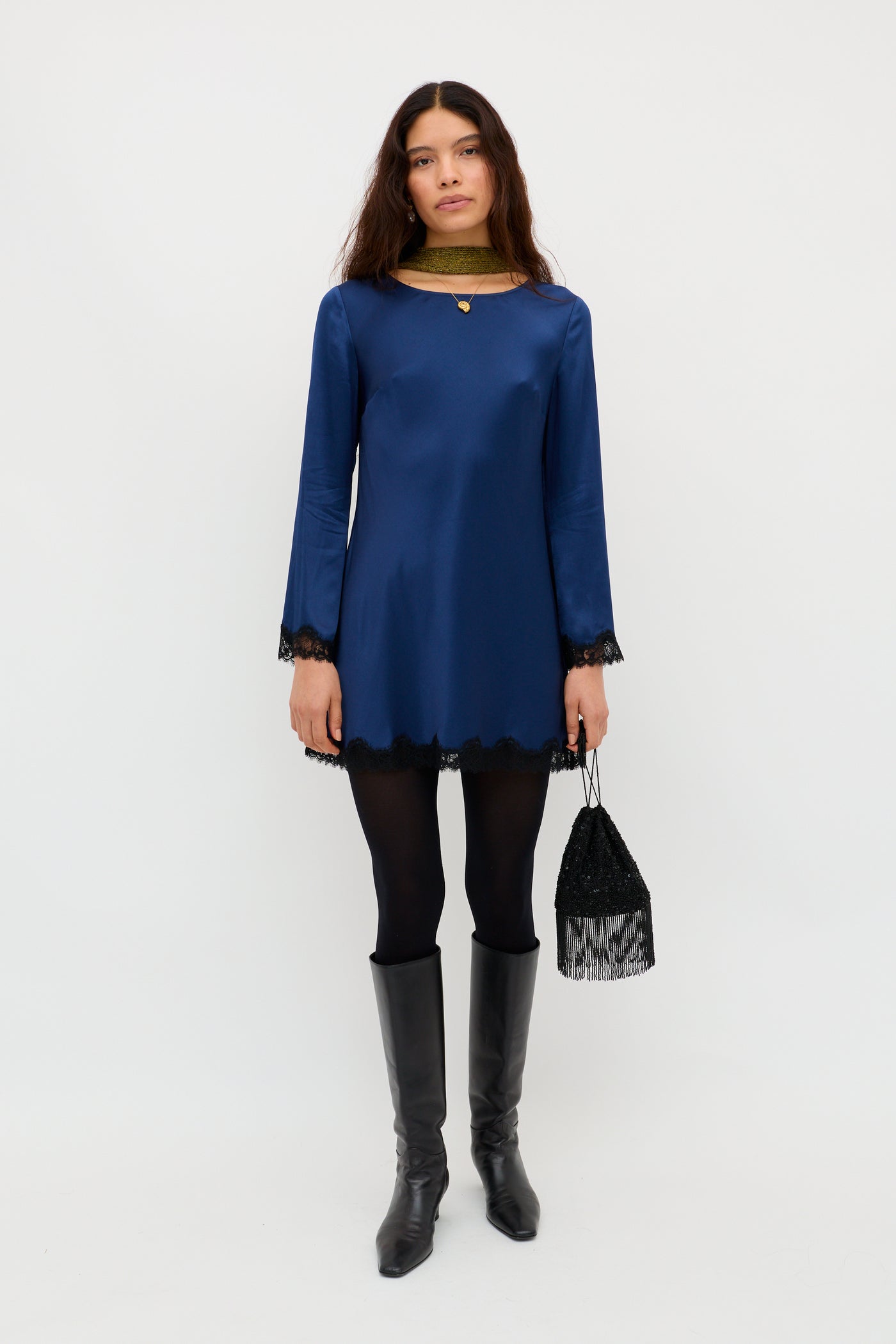 A woman stands against a white background wearing the Ridley - Midnight Navy long-sleeve mini dress with black lace trim, paired with black tights, knee-high black boots, a small fringed bag, and loose hair.