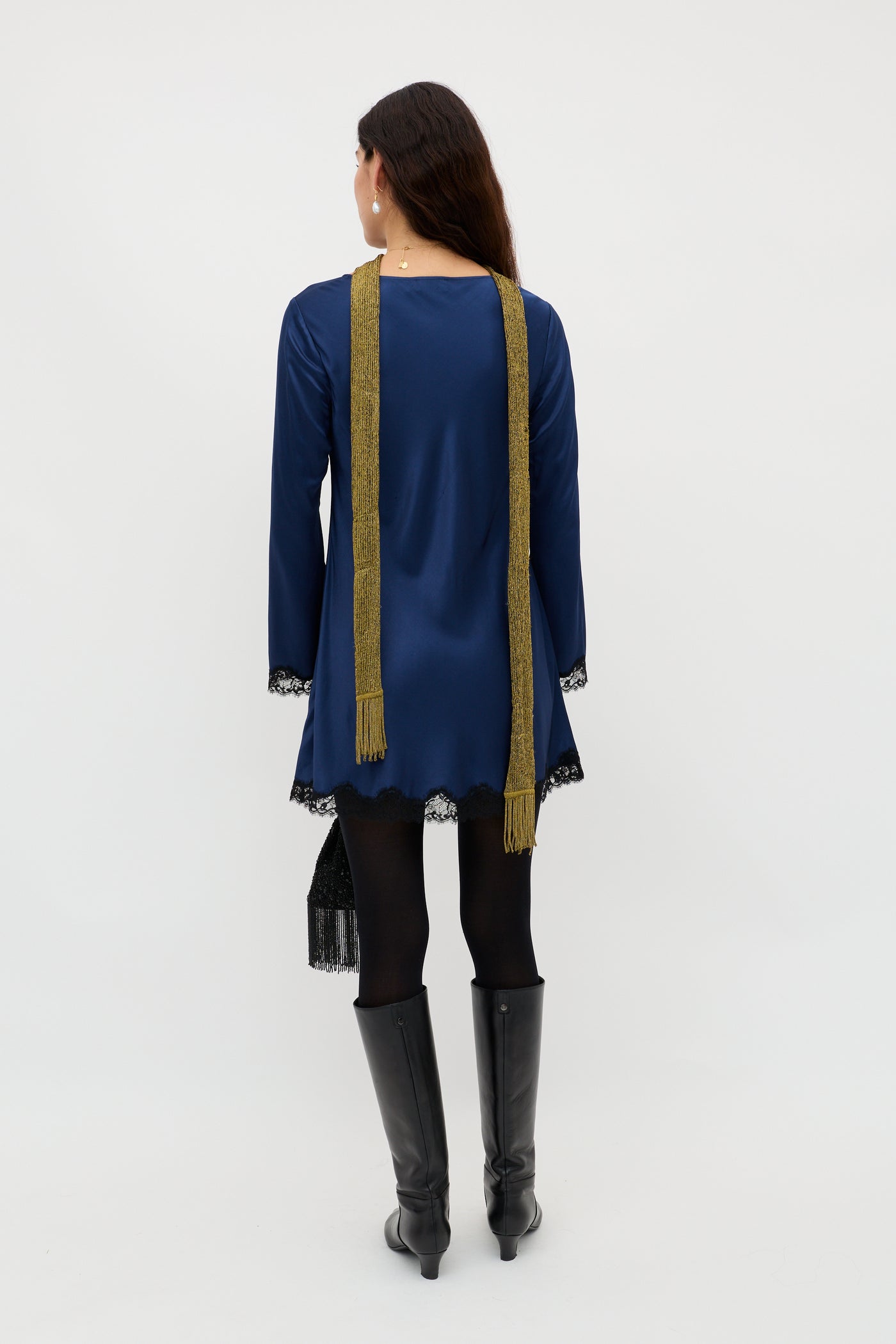 A woman faces away, wearing the Ridley - Midnight Navy long-sleeved dress with lace trim, black tights, tall black boots, a long gold scarf, and holding a small black bag against a plain white background.