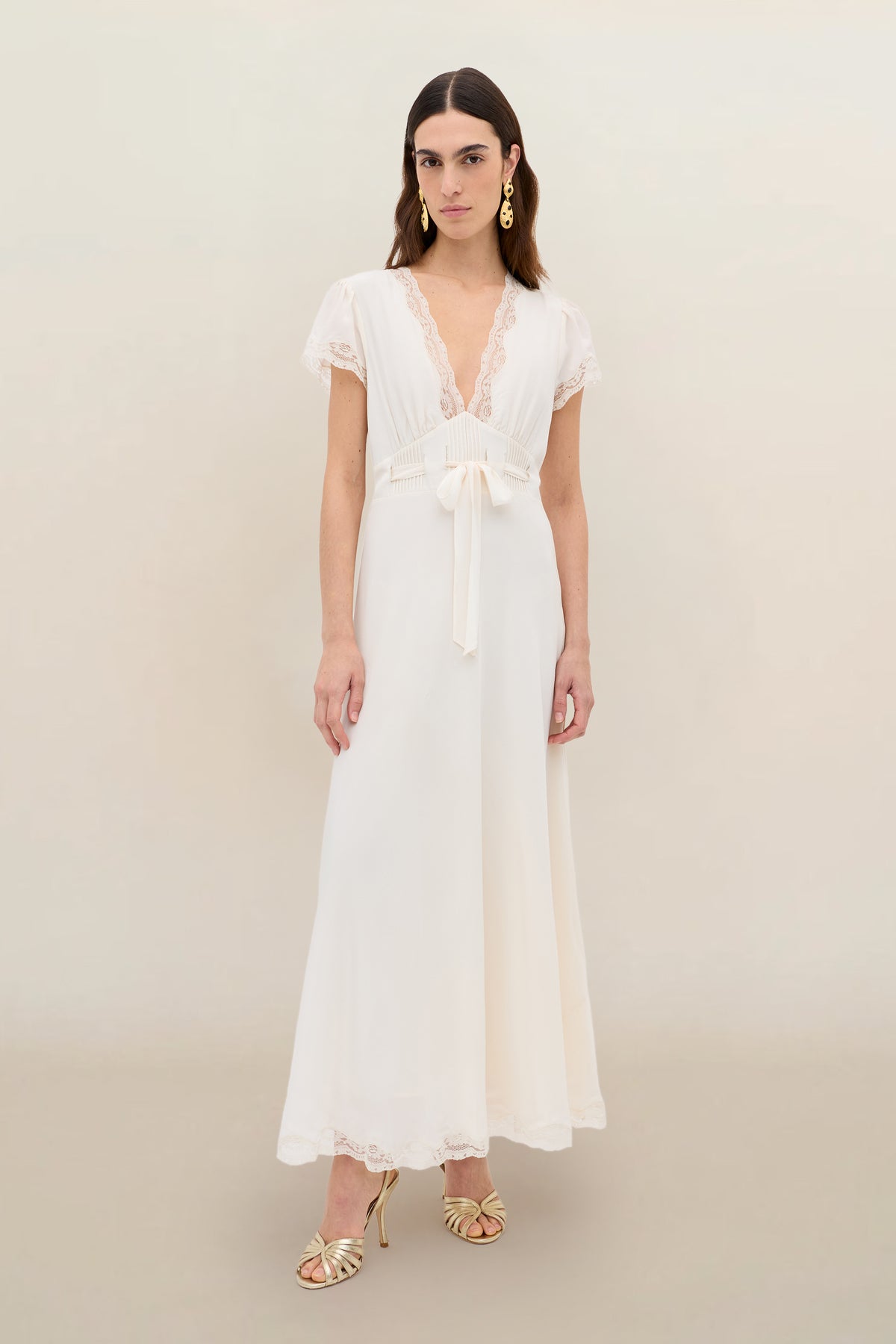 Wearing the Marlice - Pearl, a long, vintage-inspired white dress with lace trim and a deep V-neckline, she stands against a light background. Her look is finished with gold earrings, open-toe heels, and loose, center-parted hair.