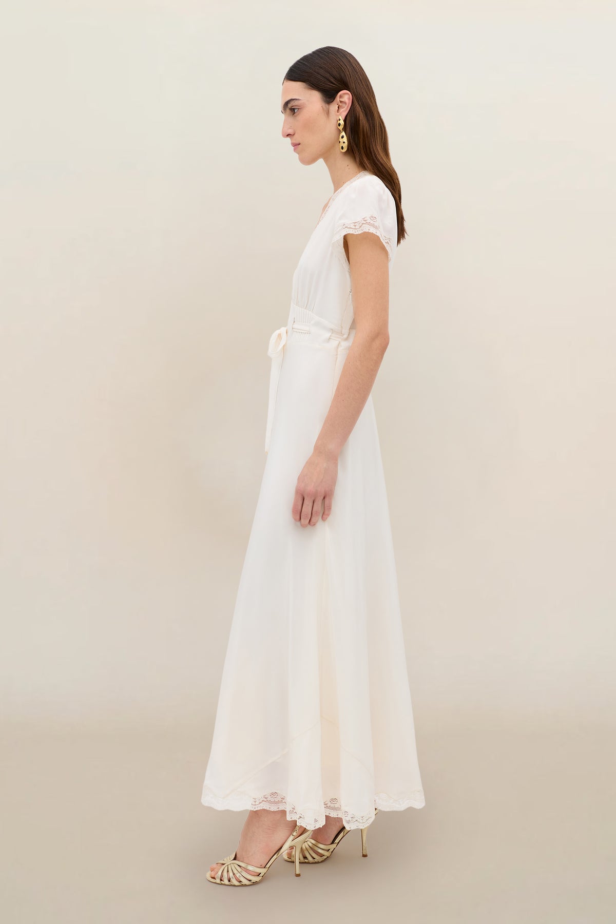 A woman stands in profile against a neutral background, wearing the Marlice - Pearl: a vintage-inspired, ankle-length ivory dress with lace trim, satin bow detail, gold earrings, and strappy heels.