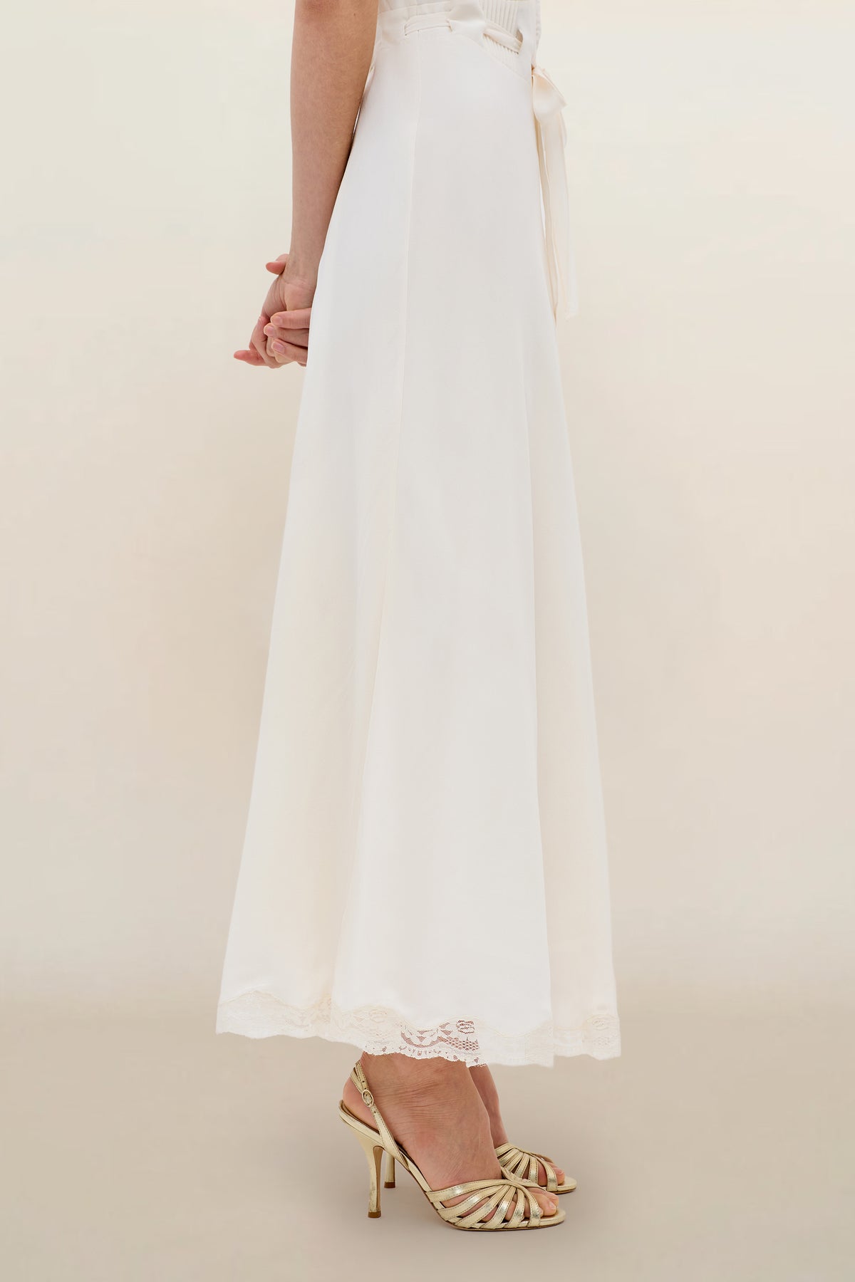 Wearing the Marlice - Pearl dress—a vintage-inspired design with a long, flowing white lace-trimmed skirt—and strappy gold heels, a person stands sideways against a simple light background.