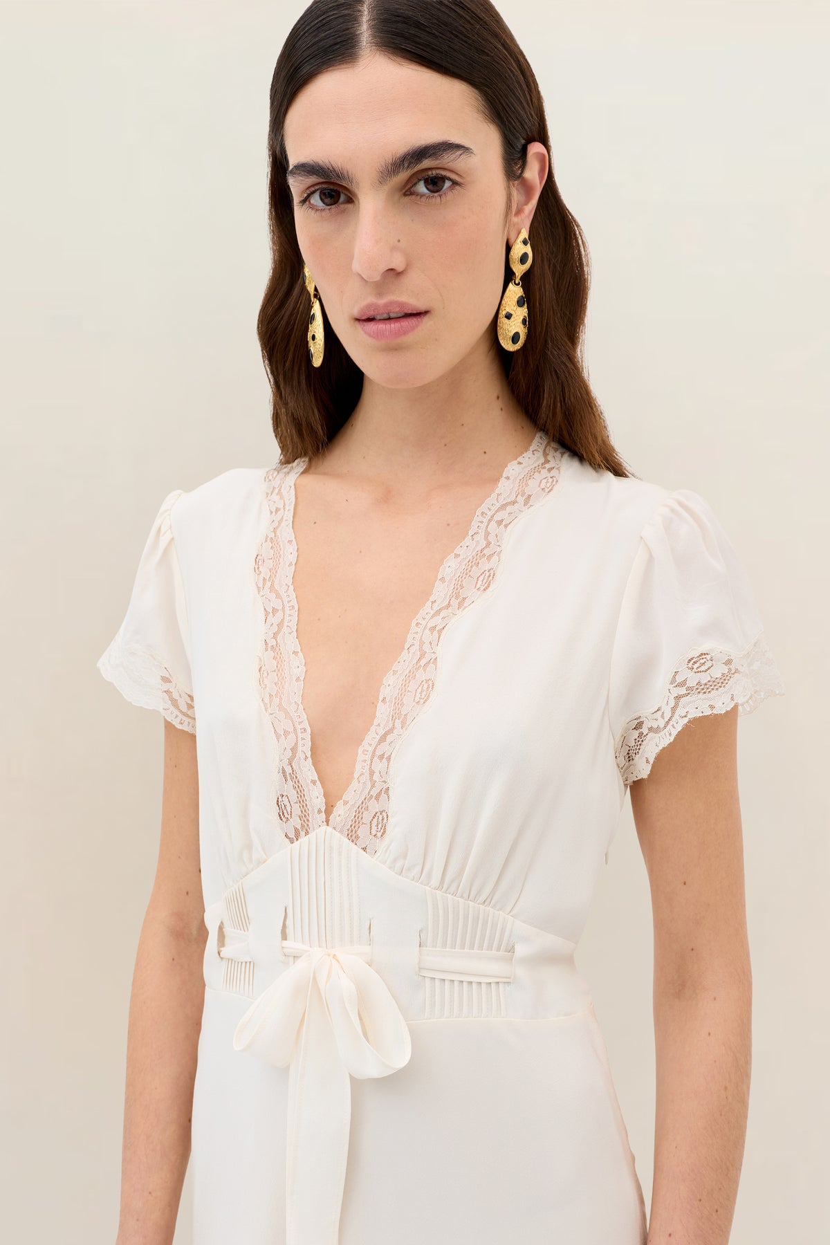 A woman with long brown hair wears gold earrings and the Marlice - Pearl dress—a cream silk midi with lace trim, short sleeves, a deep V-neckline, and a satin bow at the waist—standing against a neutral background.