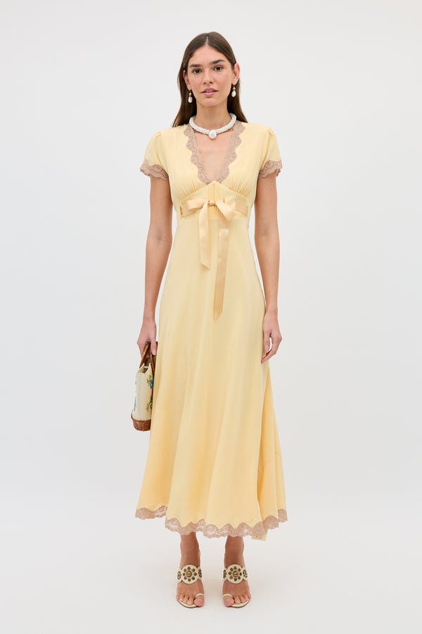A woman stands against a plain white background wearing the Marlice - Butter Yellow, a long gown with lace trims, satin waist bow, and short sleeves. She holds a small handbag and accessorizes with sandals, pearl earrings, and a necklace.