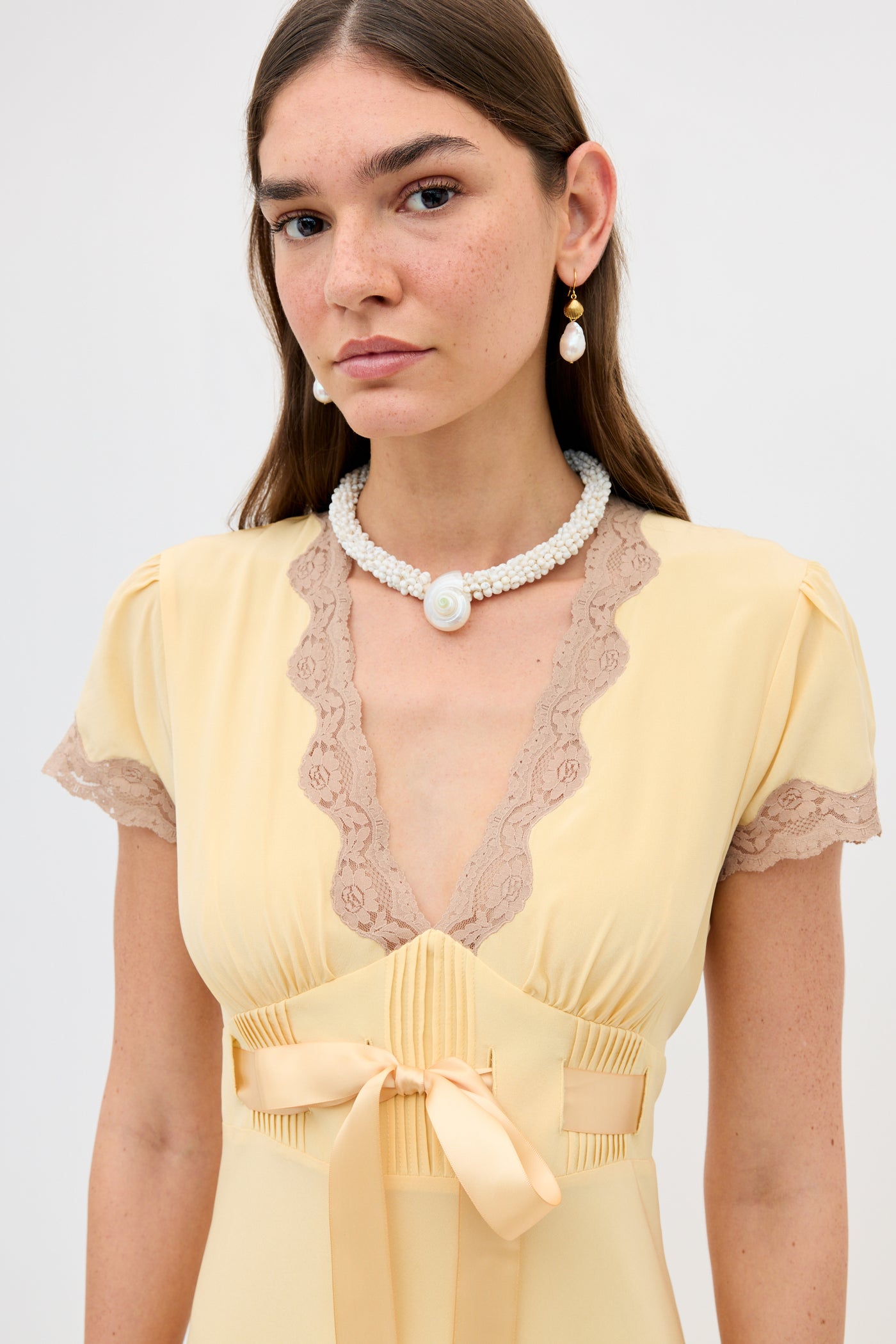 A young woman with straight brown hair wears the Marlice - Butter Yellow dress with lace trims, a satin bow, a pearl necklace with a round pendant, pearl drop earrings, and a neutral expression against a plain, light background.