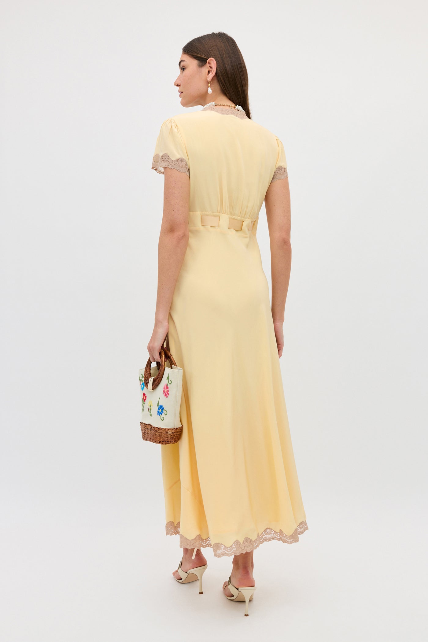 A woman stands facing away in a Marlice - Butter Yellow dress with lace trims, holding a colorful patterned handbag and wearing cream high heels. The background is plain and light.
