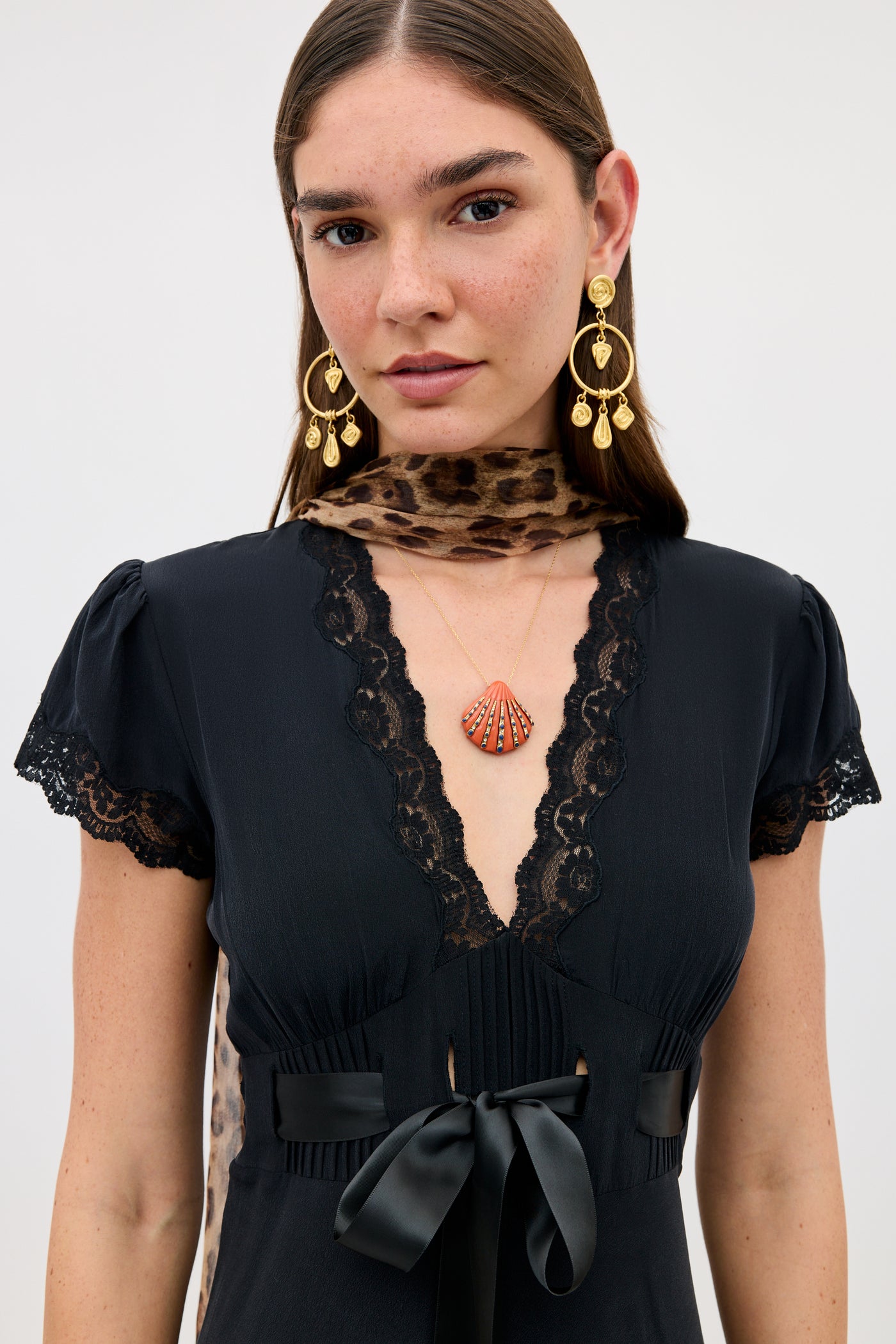 A woman with straight brown hair wears the Leanira - Orange dress, paired with gold statement earrings and a shell pendant necklace, standing against a plain white background.