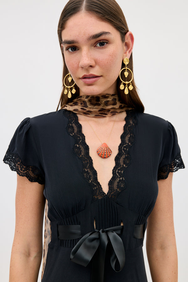 A woman with straight brown hair wears the Leanira - Orange dress, paired with gold statement earrings and a shell pendant necklace, standing against a plain white background.