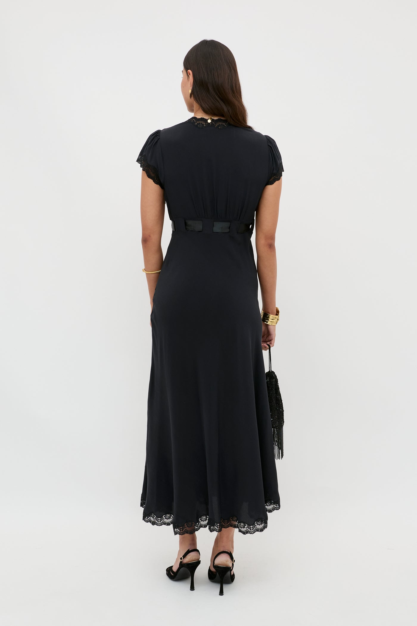 A woman stands facing away, wearing the Marlice - Black silk midi dress with lace trim. She carries a black fringe bag, wears black heels, gold bracelets, and a watch. Her dark hair is down, completing her timeless look.