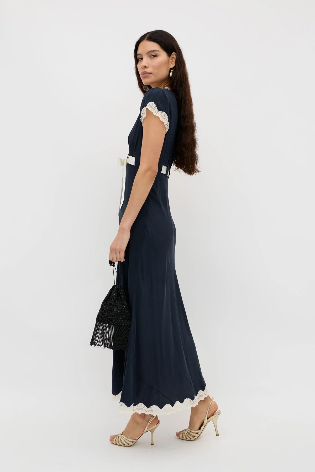 A woman with long brown hair stands sideways against a plain white background, wearing the Marlice - Deep Navy dress with white lace trim, paired with a black fringed handbag and gold heeled sandals.