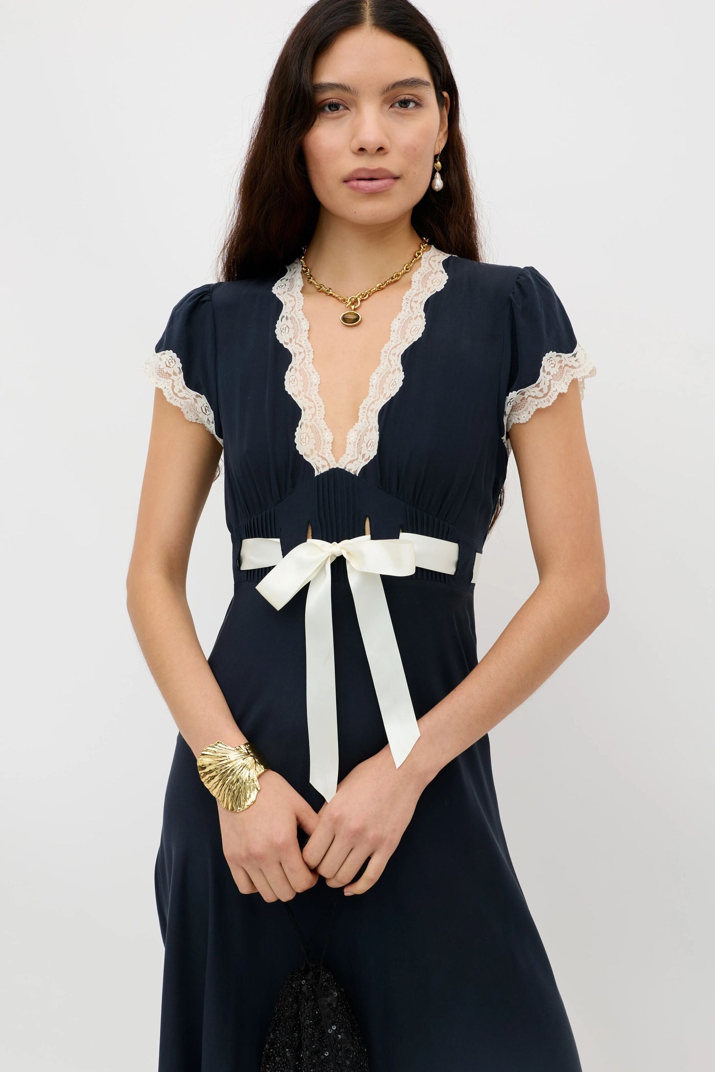 A woman in a deep navy Marlice dress with lace trims, a deep V-neckline, short sleeves, and a satin bow at the waist stands against a plain background. She accessorizes with gold shell bracelet, necklace, and earrings.