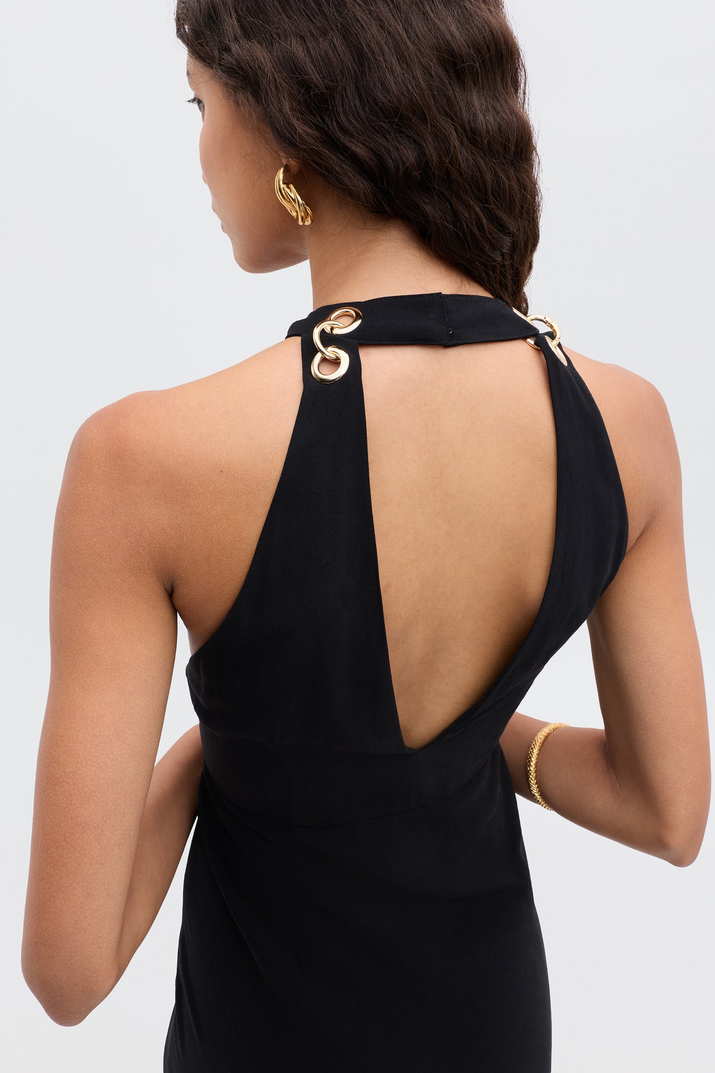 A woman with wavy brown hair wears the Eleanora - Black sleeveless dress, featuring a deep V-shaped back and gold grommet strap details. She pairs it with gold hoop earrings and a bracelet against a plain light gray background.