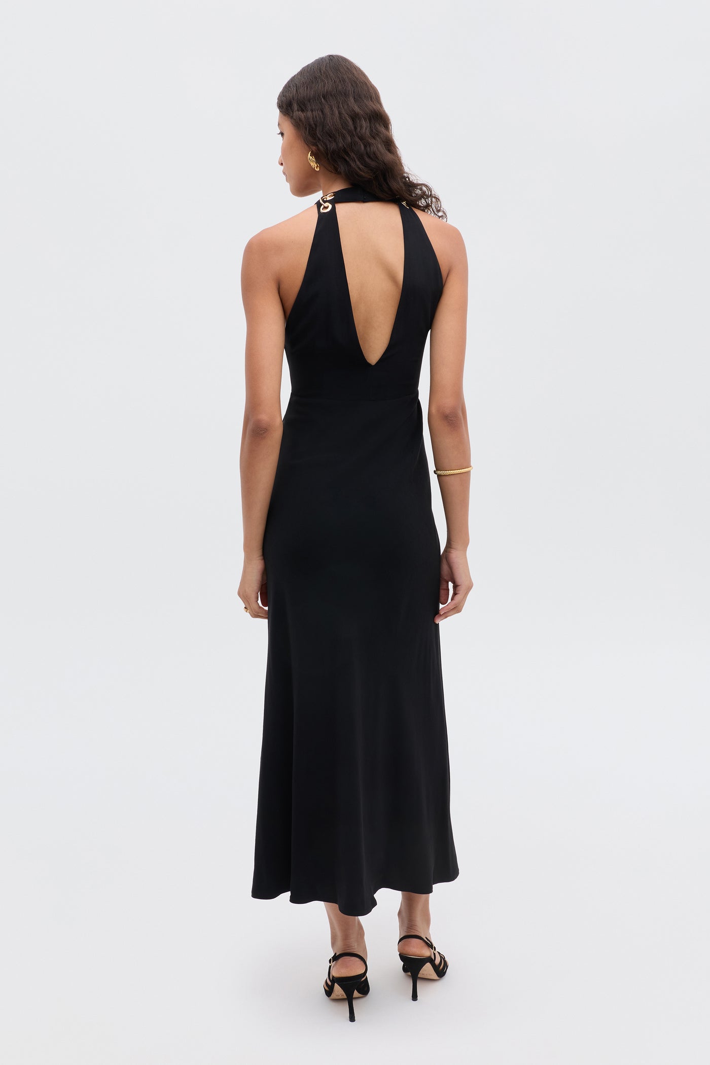 A woman with wavy hair and a gold bracelet stands facing away in the Eleanora - Black sleeveless halter-neck dress with a keyhole back and black heeled sandals, set against a plain light background.