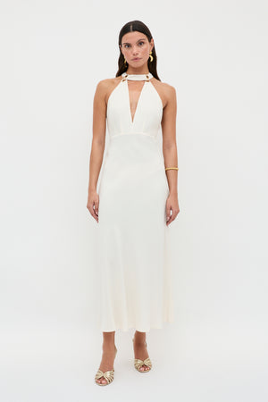 A woman stands against a white background wearing the Eleanora - Pearl dress by RIXO, featuring a sleeveless halter neck, deep V-neckline, and bias-cut hem. She accessorizes with gold earrings, a bracelet, and gold open-toe heels for a vintage-inspired look.