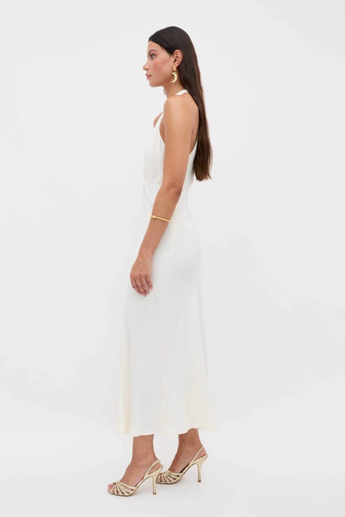A woman with long brown hair wears the Eleanora - Pearl, a vintage-inspired sleeveless RIXO dress with a bias-cut hem and heeled sandals. She accessorizes with gold earrings and bracelet, standing sideways against a plain white background.