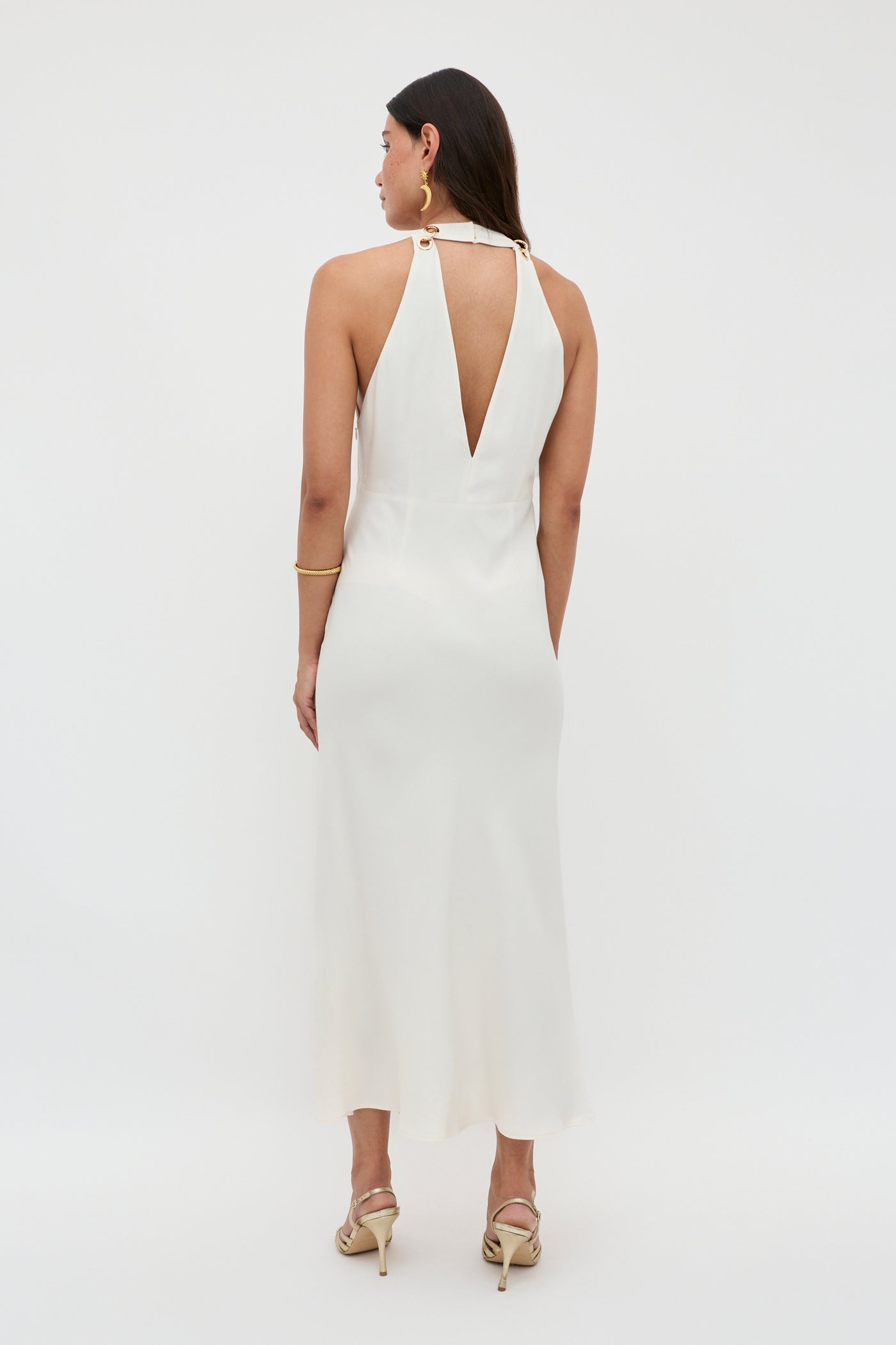 A woman with long dark hair stands facing away, wearing the Eleanora - Pearl, a sleeveless white RIXO dress with a halter neckline, bias-cut hem, and deep V-back. She accessorizes with gold hoop earrings, bangles, and strappy heeled sandals.