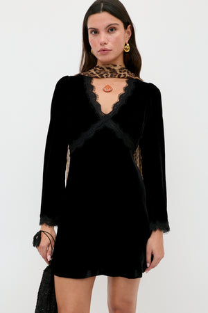 A woman stands against a white background wearing the RIXO Macie - Black dress with lace trim and long sleeves, accessorized with gold earrings, a leopard print scarf, and holding a black purse.
