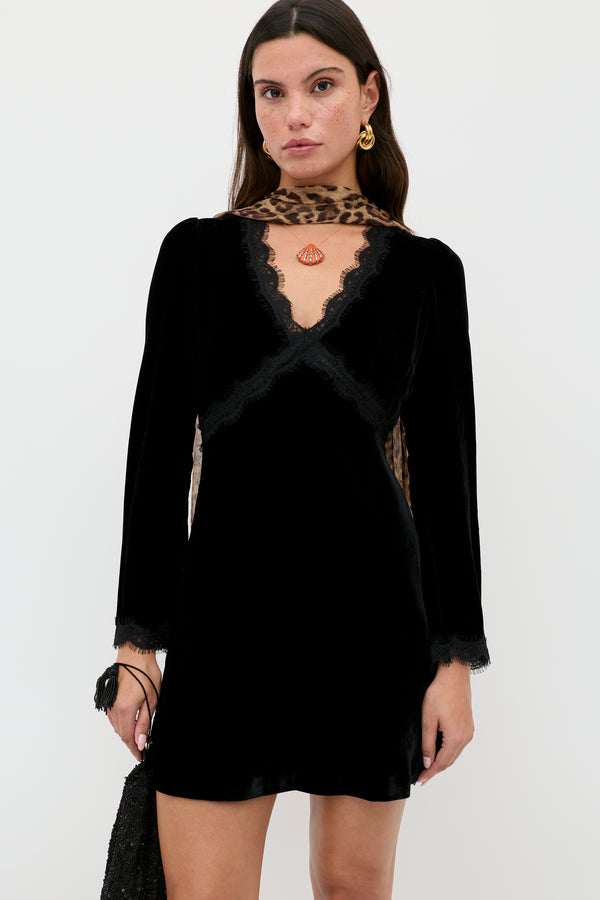 A woman stands against a white background wearing the RIXO Macie - Black dress with lace trim and long sleeves, accessorized with gold earrings, a leopard print scarf, and holding a black purse.