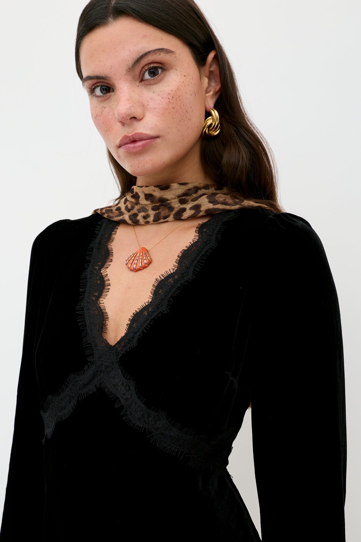 A woman with long brown hair and freckles wears a black velvet Macie dress with lace trim, a leopard print scarf, gold earrings, and a small orange seashell pendant necklace against a plain background.