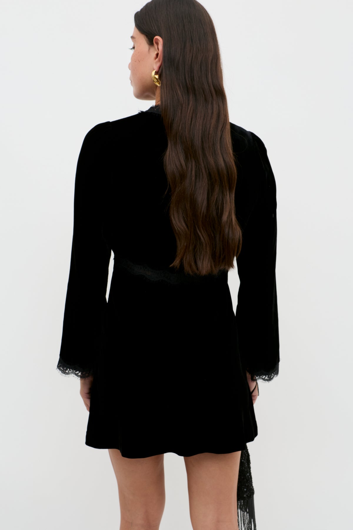 A woman with long, wavy brown hair is seen from behind wearing the Macie - Black dress with lace cuffs and gold earrings, standing against a plain white background.