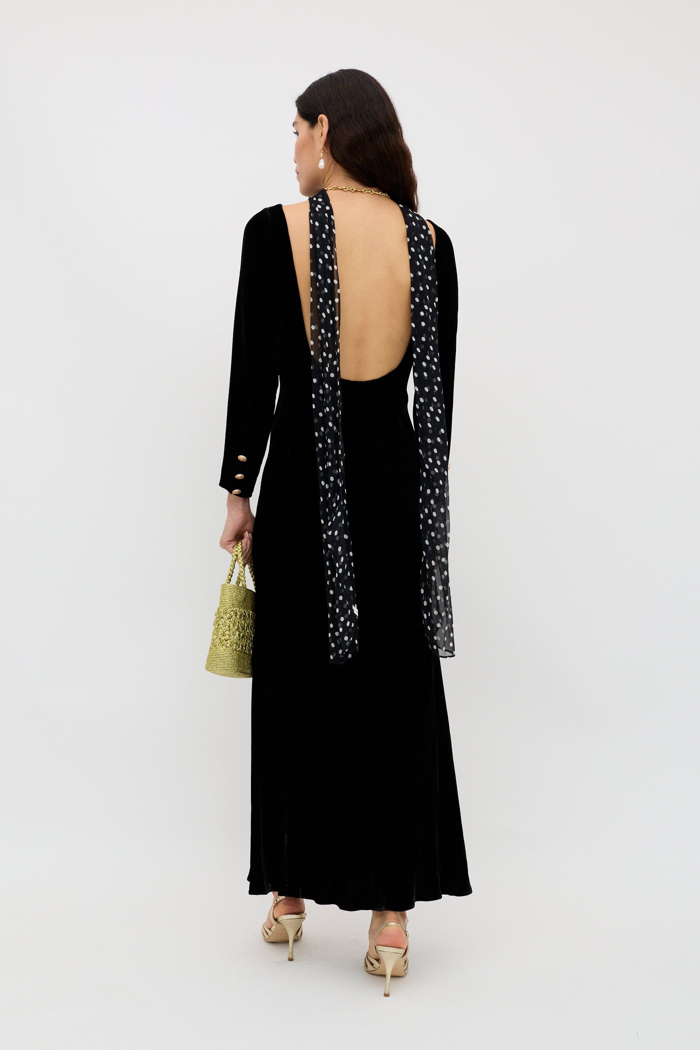 A woman in an elegant evening dress with a low-cut open back and scarf detail stands facing away, holding the Devina - Black woven handbag and wearing strappy heels against a plain white background.