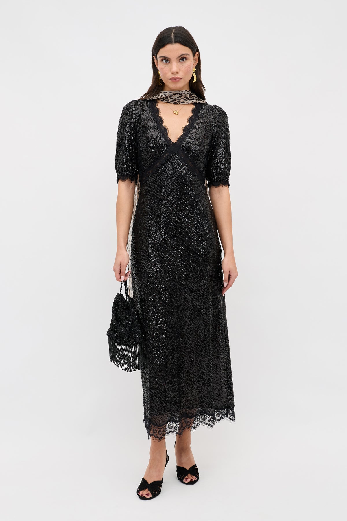 A woman wearing the Verena - Embellishment Black dress, a sparkly black lace midi with short sleeves and a deep V neckline, holds a small black handbag and wears black open-toe heels while standing against a plain white background.