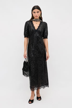 A woman wearing the Verena - Embellishment Black dress, a sparkly black lace midi with short sleeves and a deep V neckline, holds a small black handbag and wears black open-toe heels while standing against a plain white background.