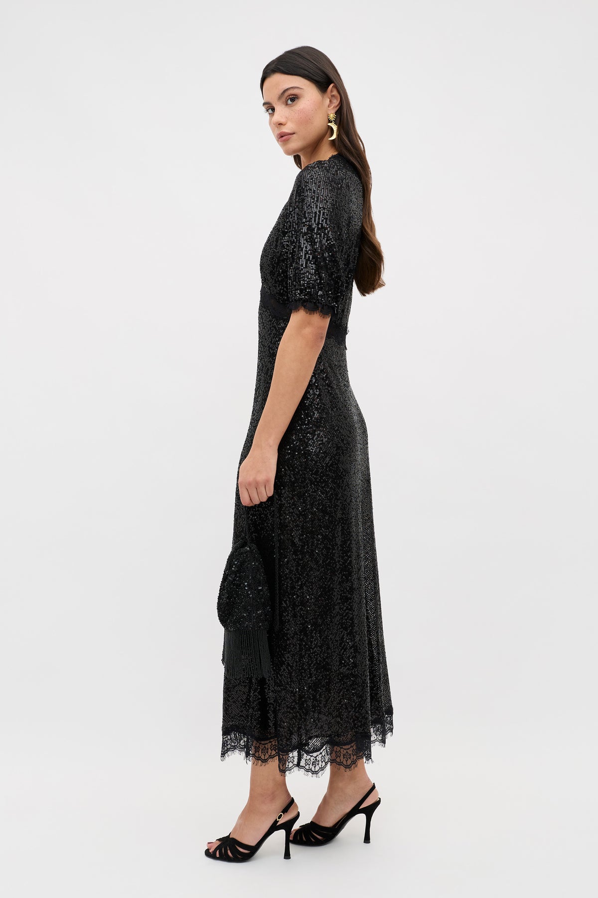 A woman stands against a plain white background wearing the Verena - Embellishment Black dress, featuring black sequins, short sleeves, and an ankle-length silhouette with a lace hem. She holds a matching clutch, wears black heels and gold earrings, and her long brown hair cascades down her shoulders.