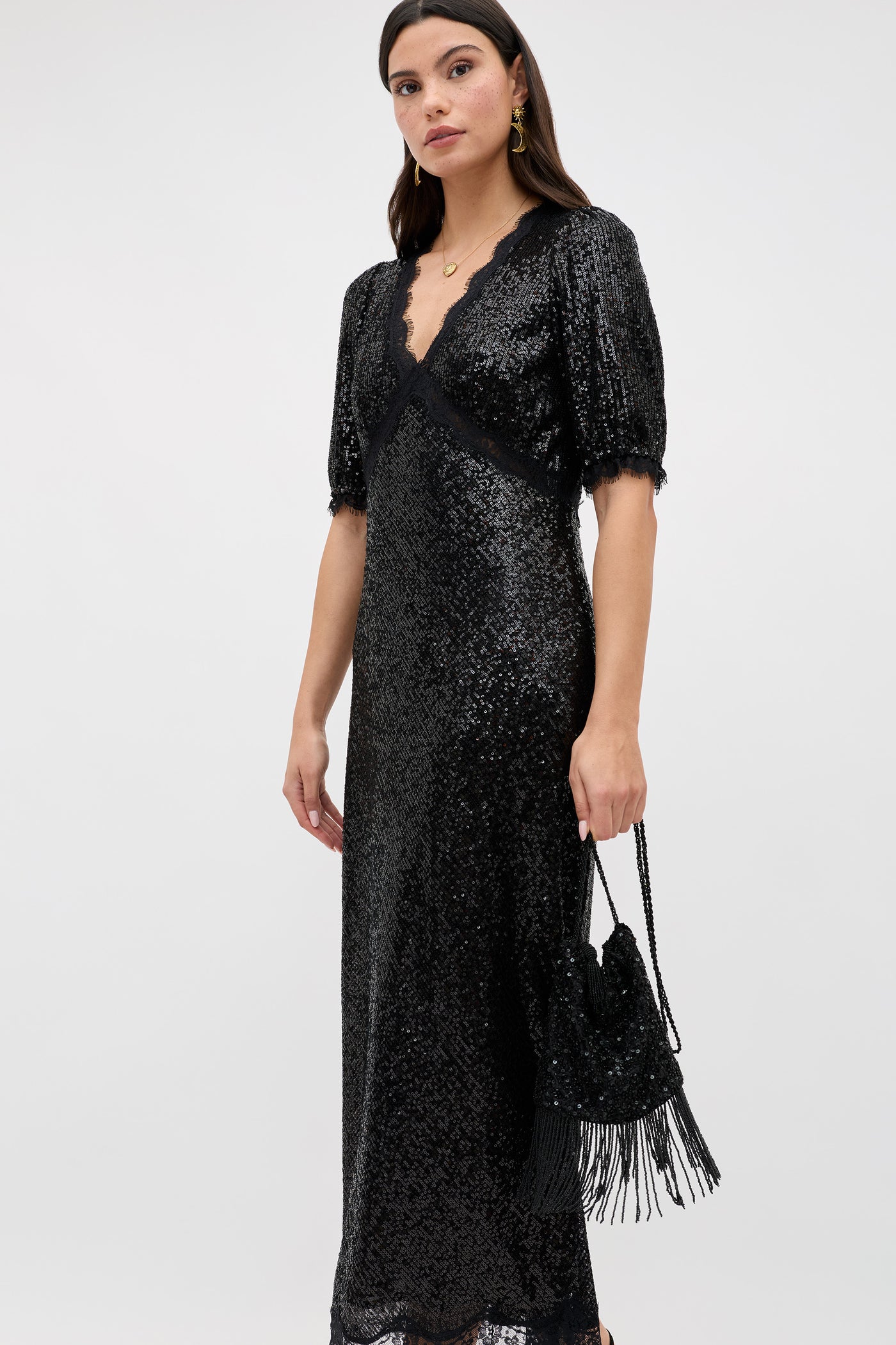 Wearing the Verena - Embellishment Black, a woman stands against a plain light background, holding a matching black fringed purse and gazing neutrally at the camera.