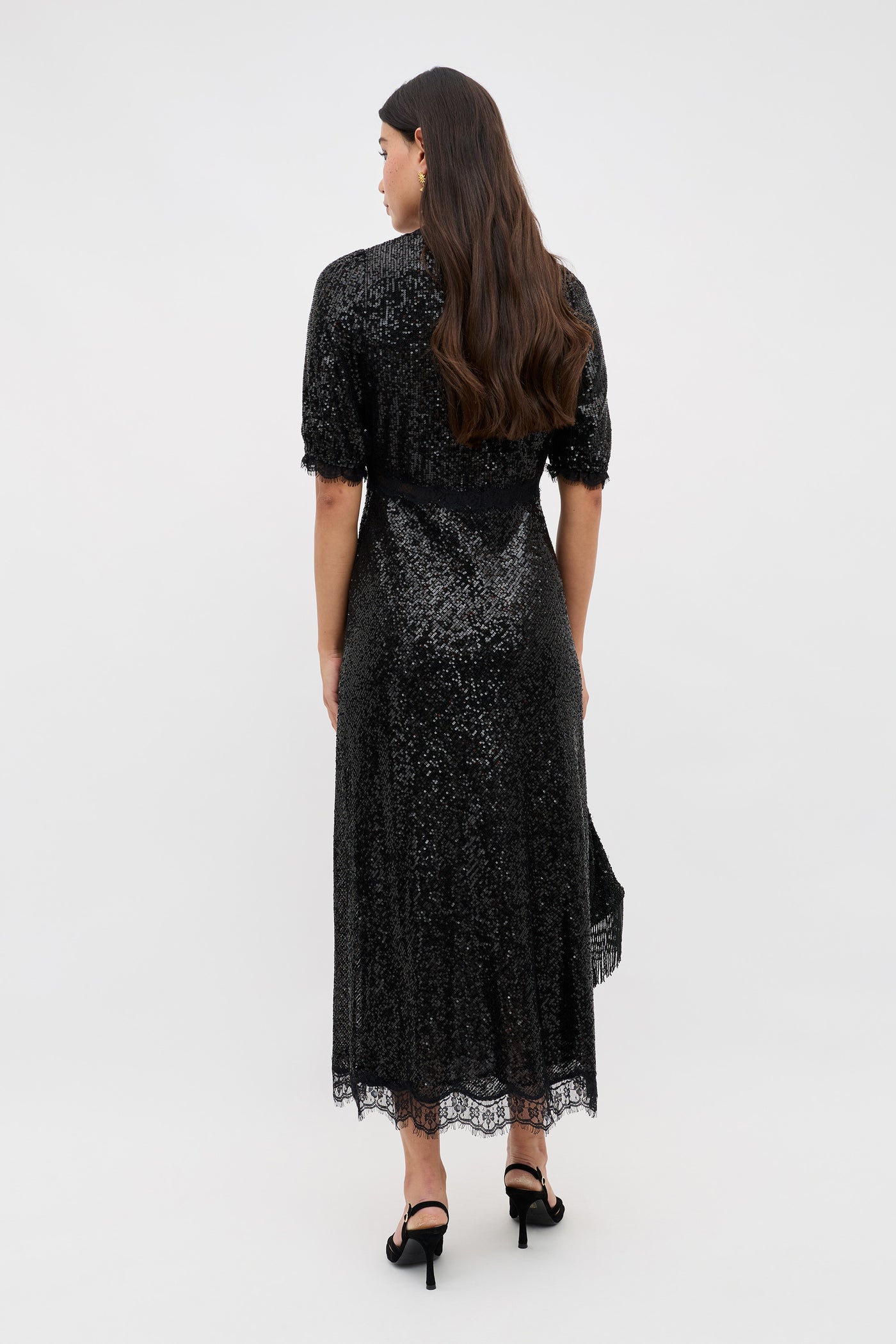 A woman with long brown hair stands facing away, wearing the Verena - Embellishment Black sequined midi dress with lace trim and black high-heeled shoes against a plain light background.