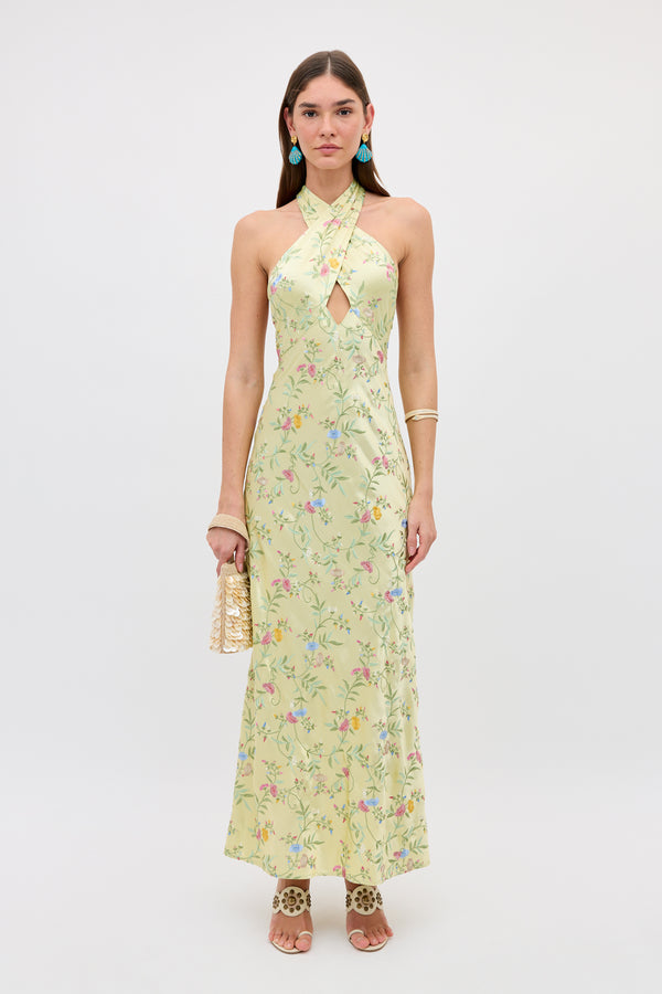 A woman in the Dune - Botanical Embroidery Green halterneck dress holds a gold purse. She has straight brown hair, turquoise earrings, gold bracelets, and leopard-print heeled sandals against a plain white background.