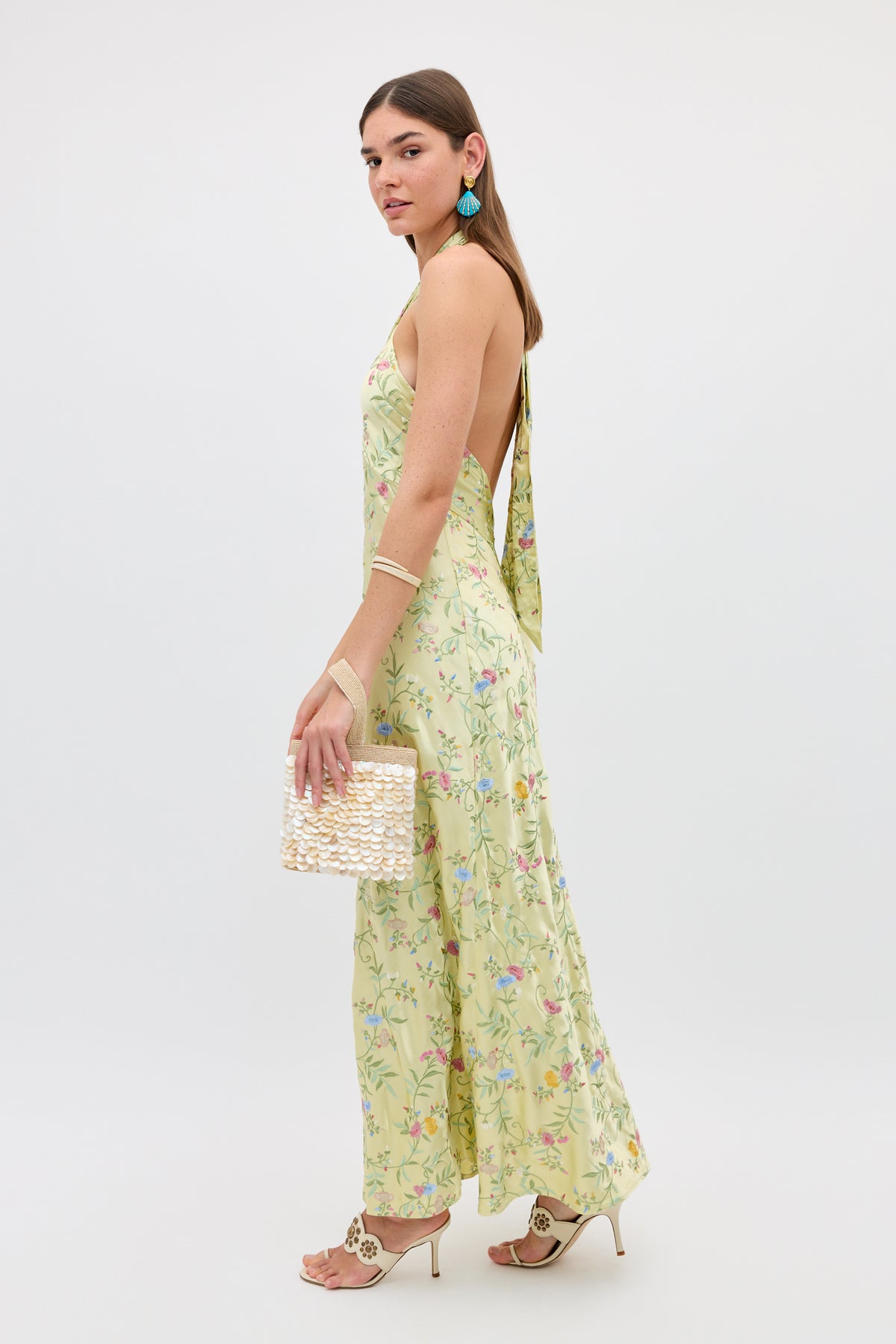 A woman poses in the Dune - Botanical Embroidery Green halterneck dress with wide legs, styled with beige heels, turquoise earrings, bracelets, and a woven clutch. Her hair is down as she stands against a plain white background.